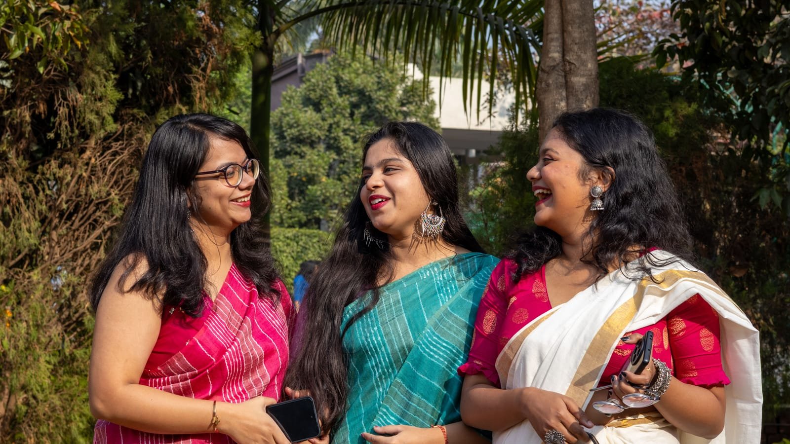 Three women friends in colourful saris laughing together in a leafy garden