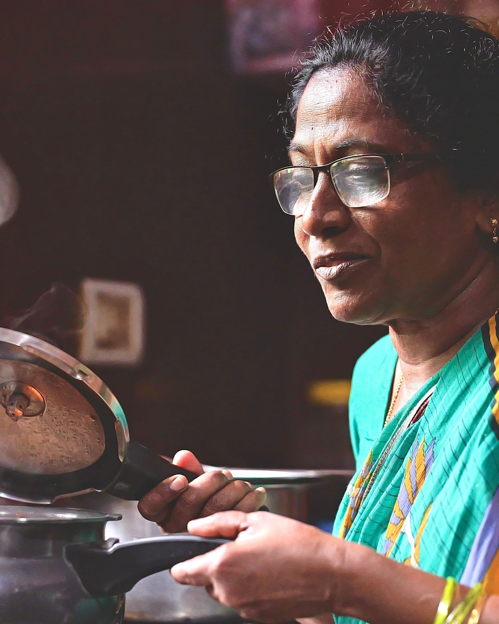 A woman in a green and orange sari lifts the lid off a steaming pressure cooker in her home kitchen
