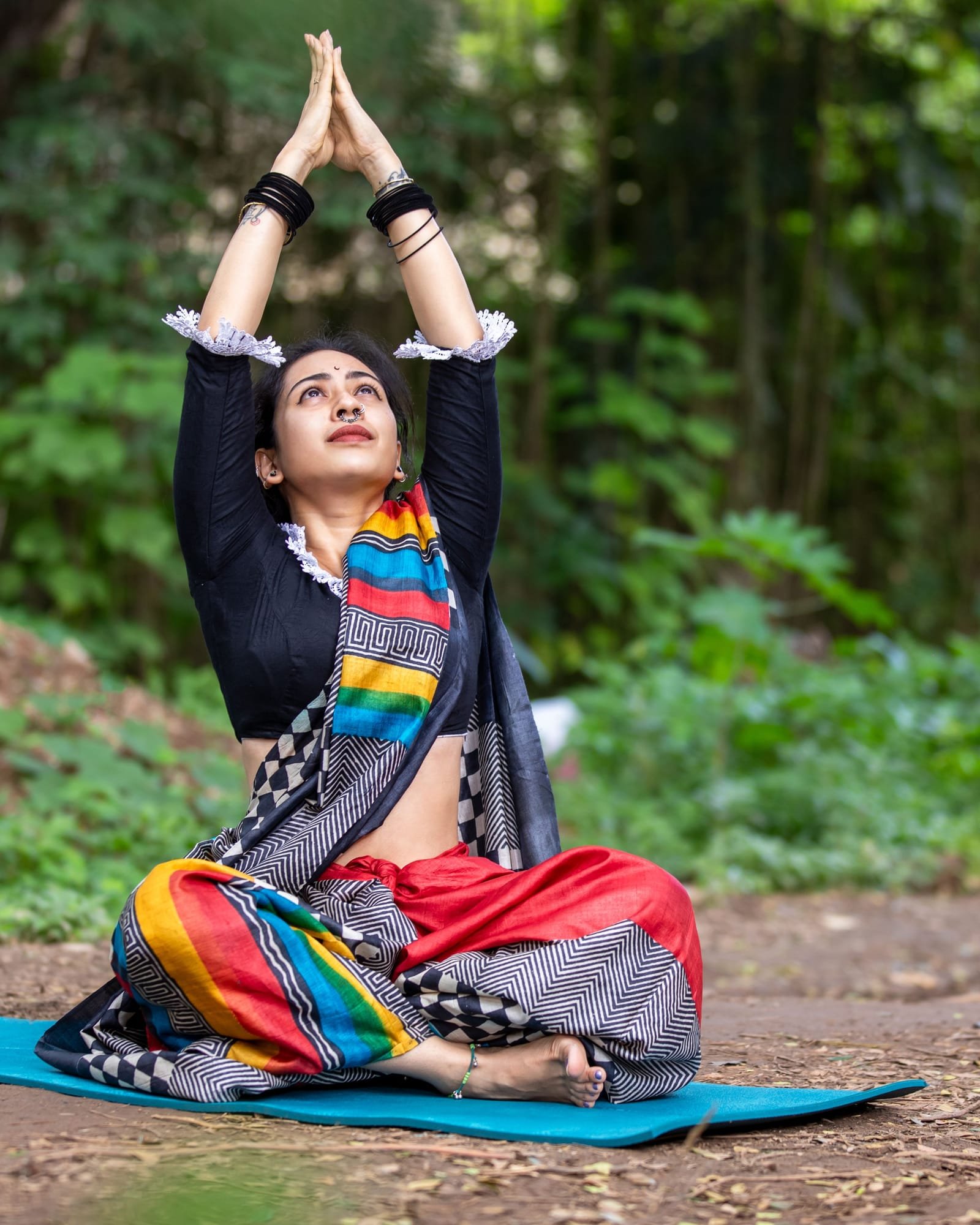 A young woman in a striped sari sits cross-legged on a yoga mat outdoors, arms stretched overhead