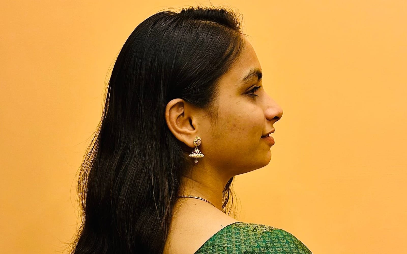 Side profile of a young Indian woman in a green sari and gold jhumka earrings, her neck visible against a warm yellow wall.