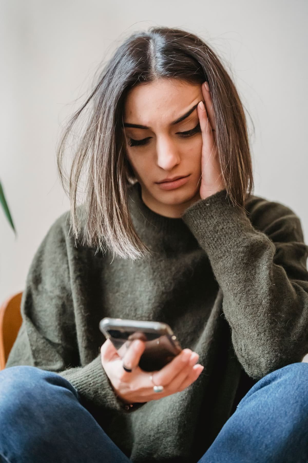 A woman sits holding her phone, one hand pressed to her forehead