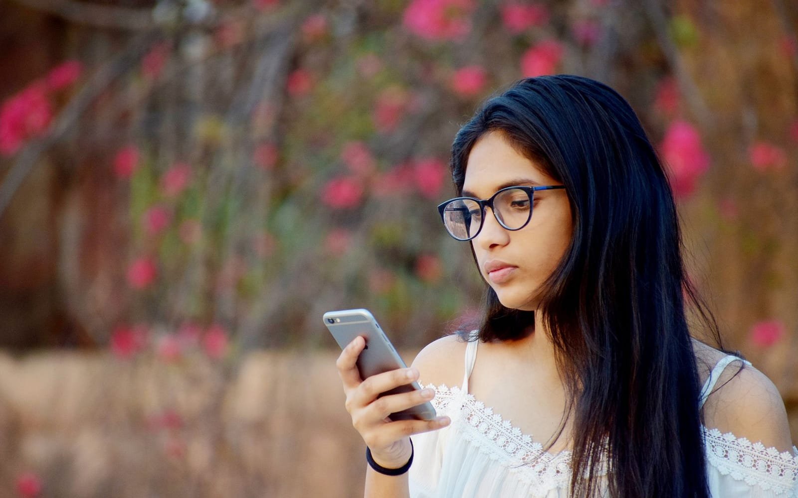 A young Indian woman in glasses looks thoughtfully at her phone, with blurred pink bougainvillea behind her.