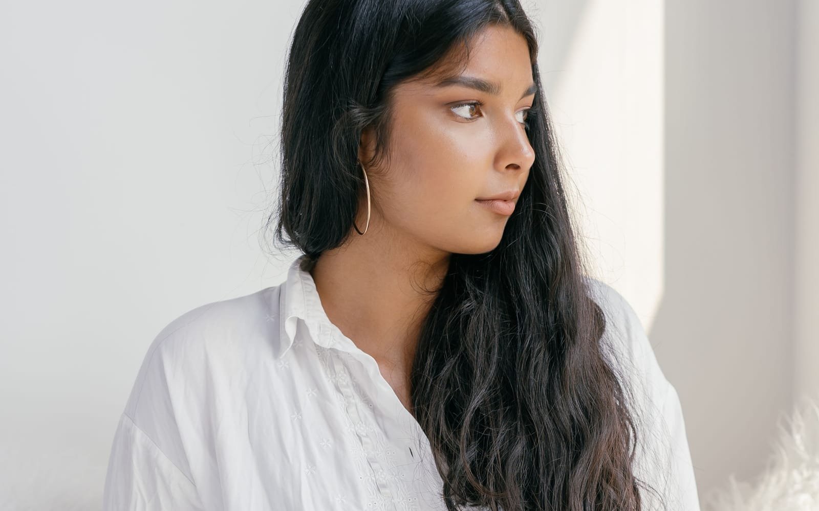 A young South Asian woman in a white shirt pauses and gazes toward a bright window.