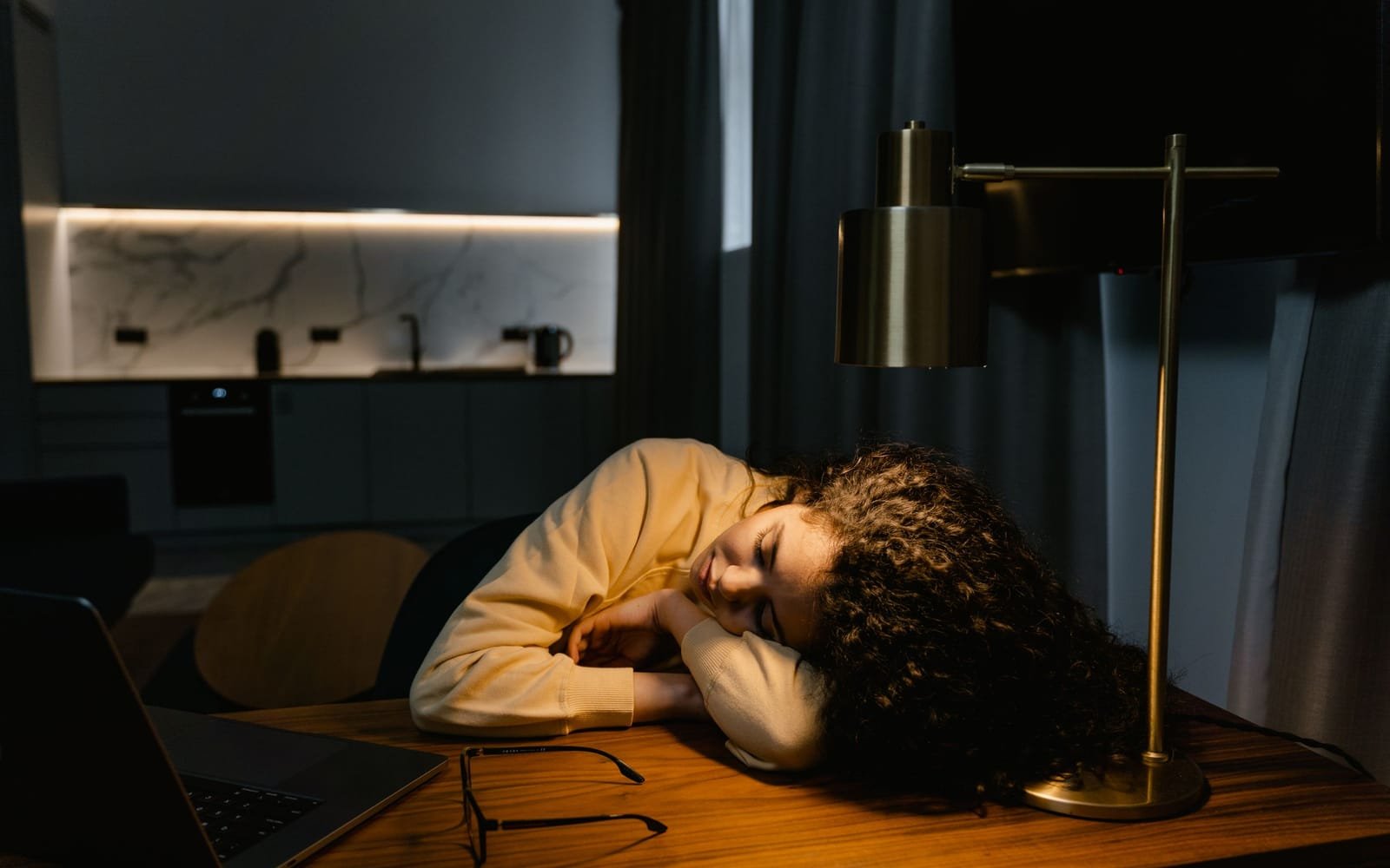 A woman rests her head on her arms at a desk beside an open laptop and her glasses, lit by a warm desk lamp.