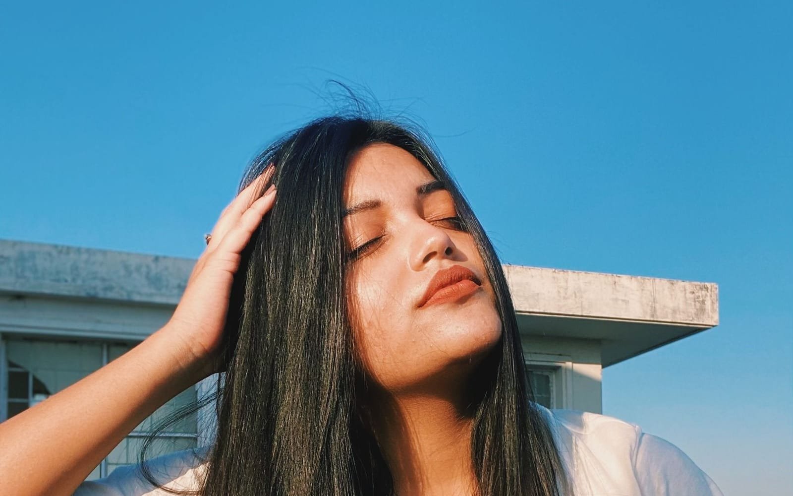 A young Indian woman runs her fingers through her long dark hair, eyes closed, in afternoon sun.
