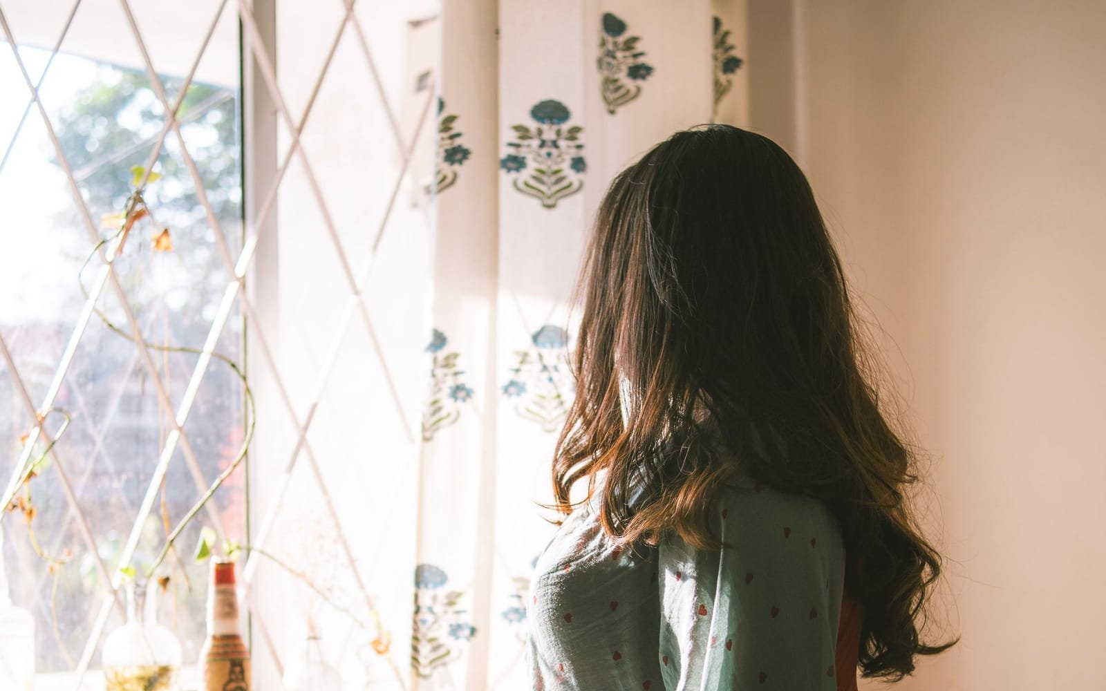 A woman with long dark hair stands at a sunlit window with block-print curtains, quietly looking out.