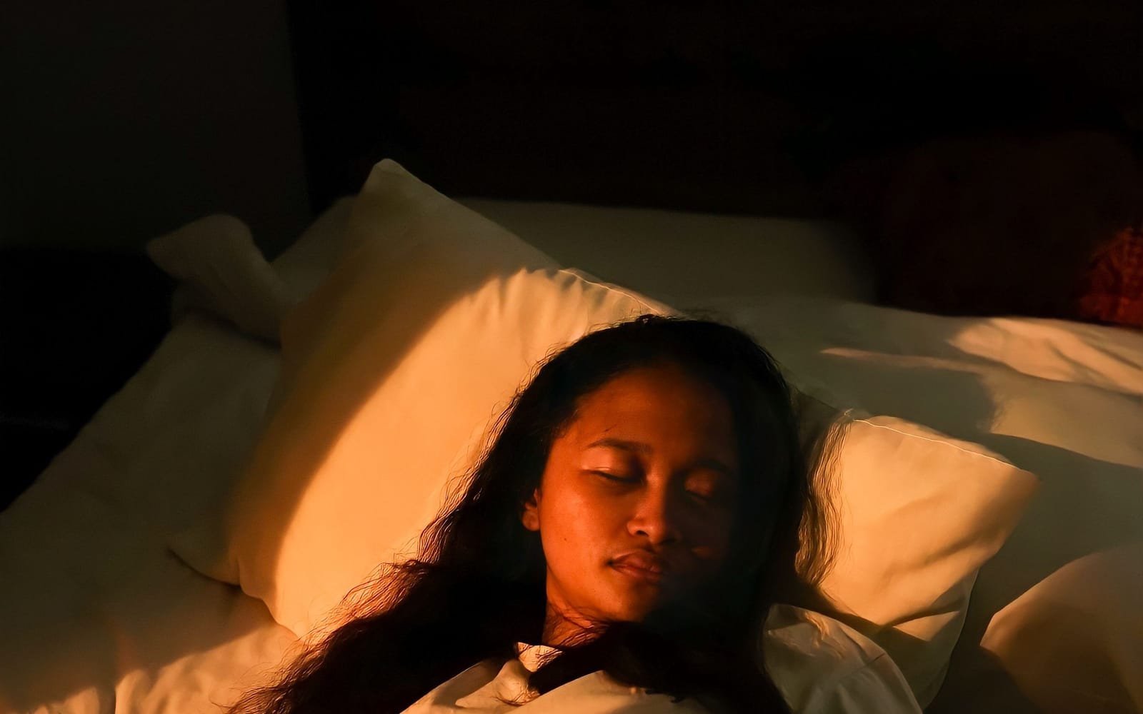 A young woman lies in bed with her eyes closed as warm lamplight falls across her pillow.