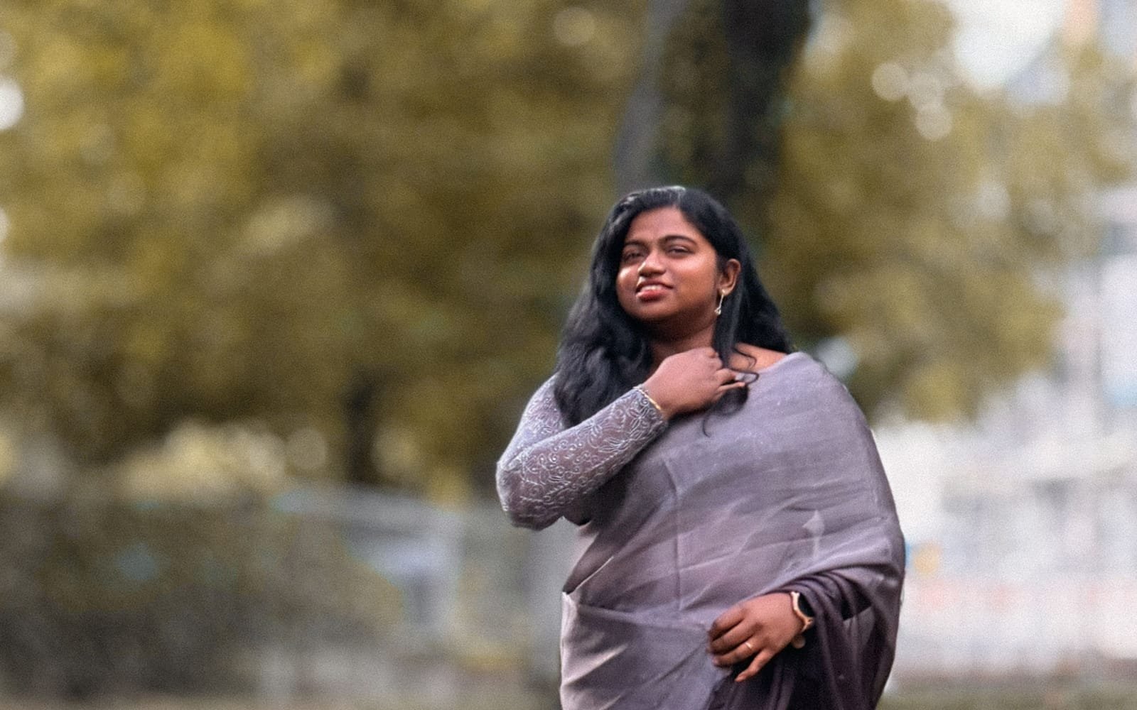 A smiling South Asian woman in a grey saree walks through a leafy park in soft daylight.