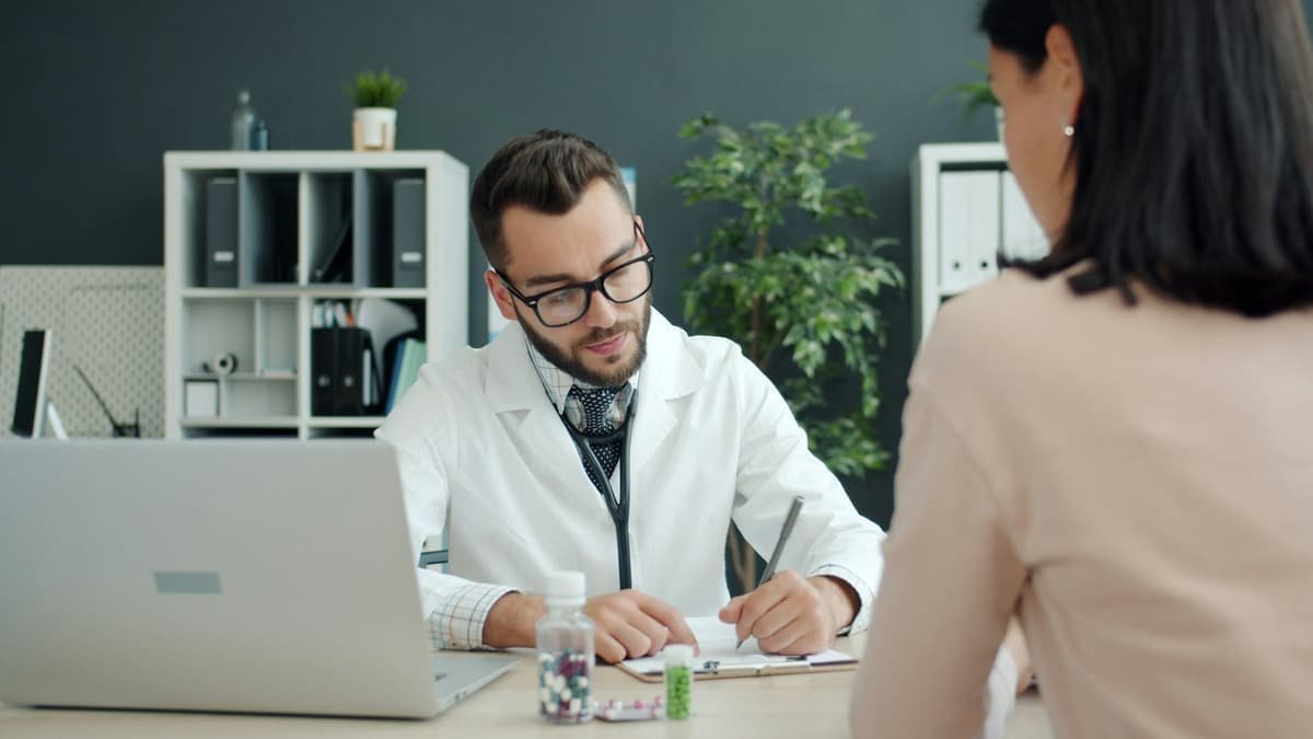 A doctor takes notes at his desk while a woman explains her history