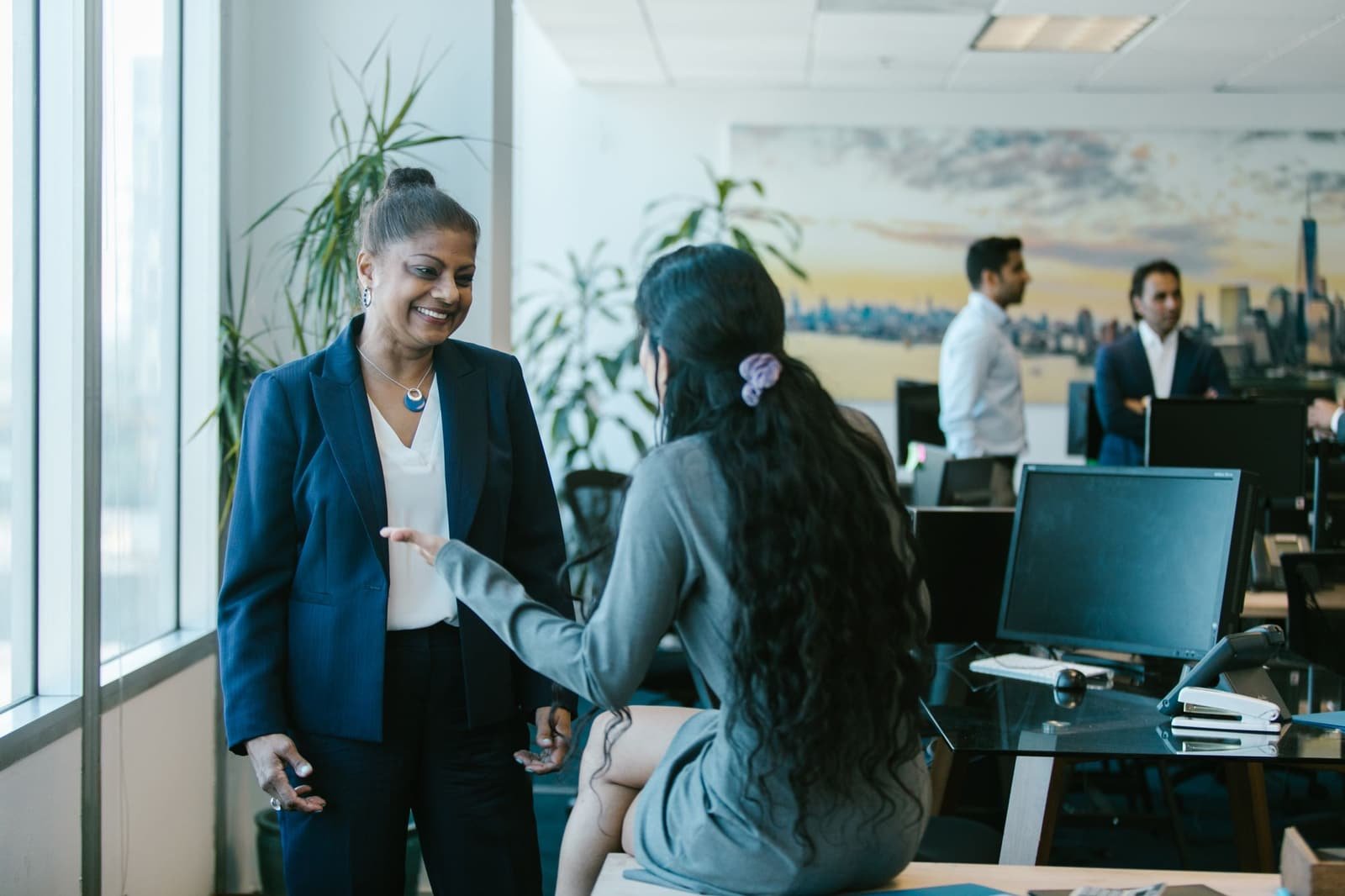 Two Indian women colleagues talking at a desk in a bright, modern office