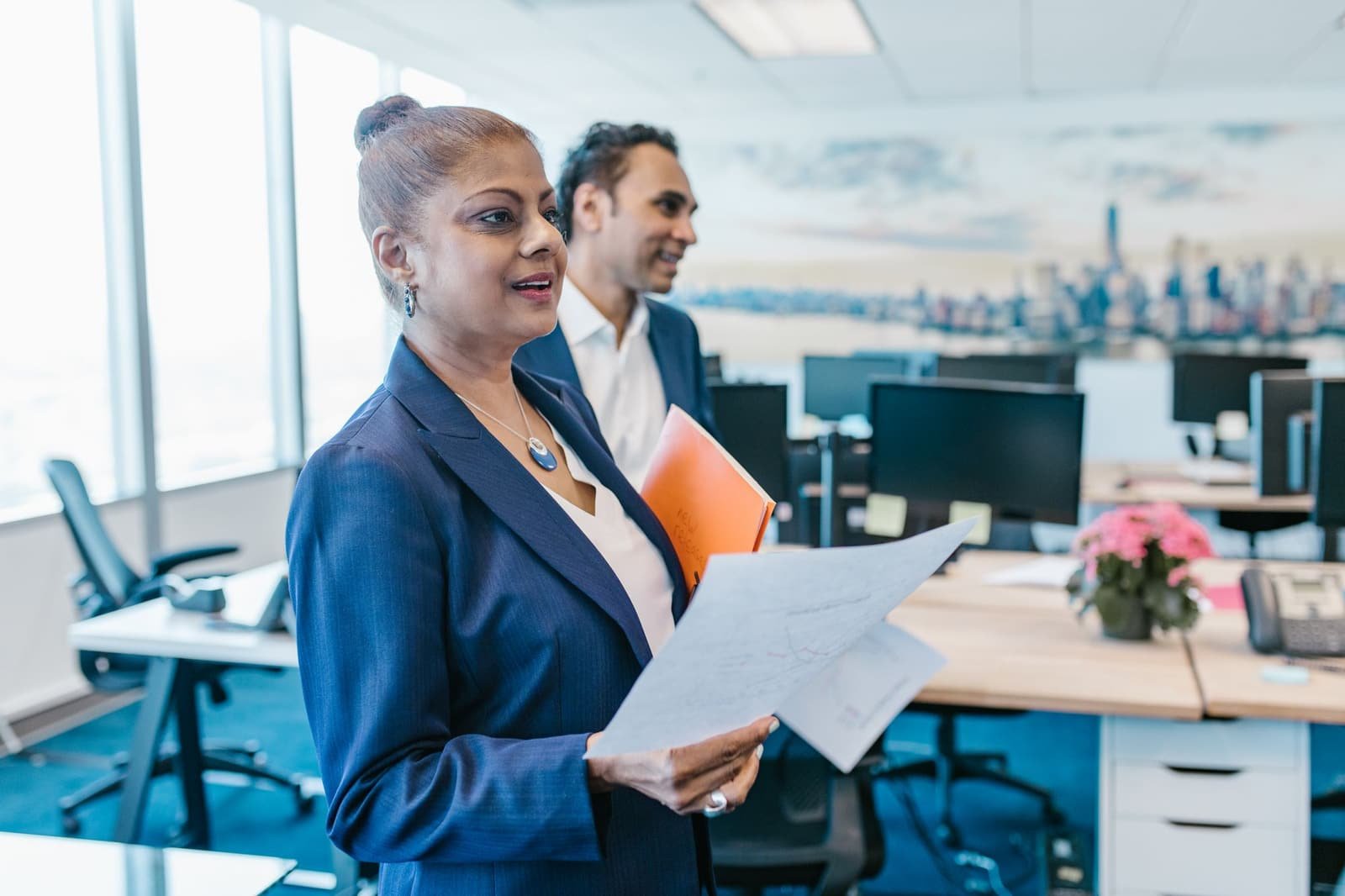An Indian woman manager in a navy blazer reading papers with a colleague in an open-plan office