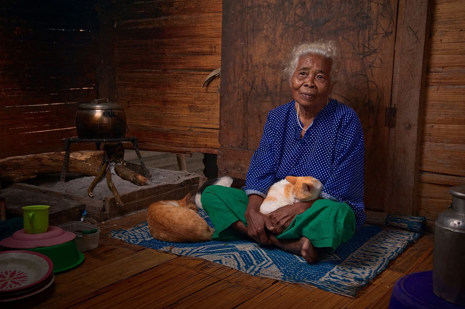 An elderly woman in a blue blouse sitting on a mat in her wooden home, holding a sleeping cat