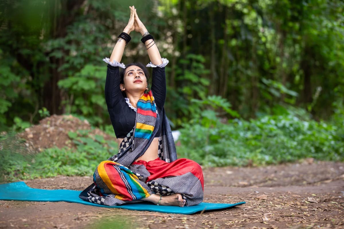 A woman sitting cross-legged on a yoga mat outdoors with her arms raised, in soft daylight under trees